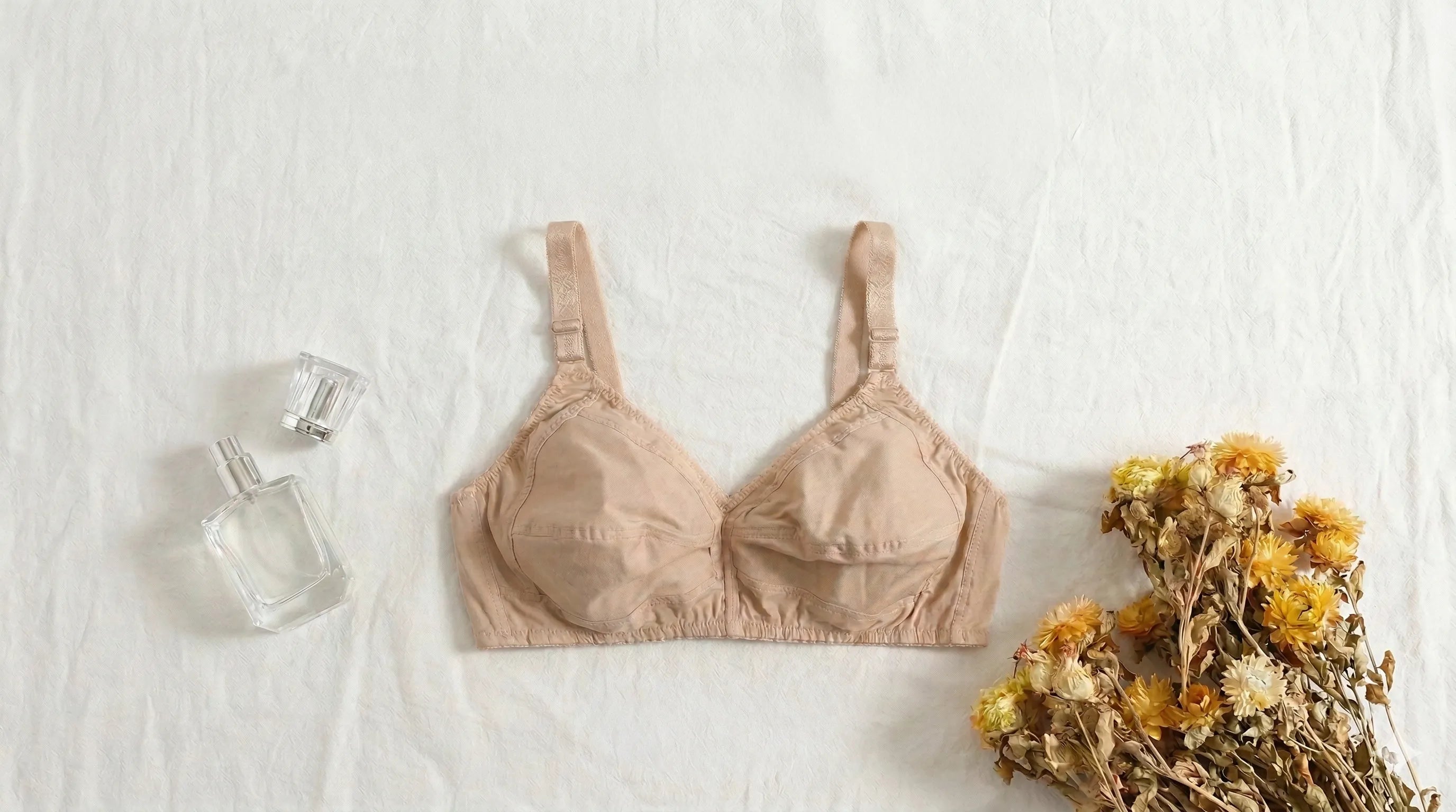 Beige bra on a white surface with a bottle of perfume and dried flowers.
